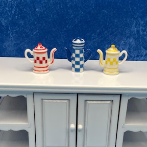 May include: Three miniature teapots in red and white, blue and white, and yellow and white checkered patterns. The teapots are displayed on a white table against a blue background. The teapots have small handles and spouts.