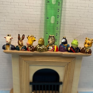 May include: A collection of miniature animal figurines, each wearing a crown and formal attire. The figurines include a goat, rabbit, giraffe, bear, wolf, bird, frog, and fox. They are displayed on a wooden shelf, with a ruler in the background.