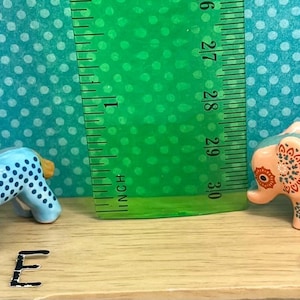 May include: Two small figurines: a blue and white spotted horse with an orange mane and a pink elephant with a decorative floral pattern. A green ruler is positioned between them, measuring in inches.
