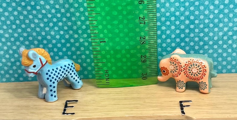 May include: Two small figurines: a blue and white spotted horse with an orange mane and a pink elephant with a decorative floral pattern. A green ruler is positioned between them, measuring in inches.