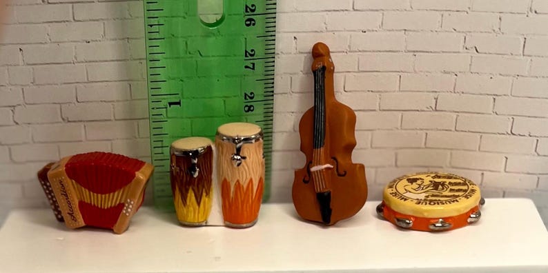 May include: Miniature musical instruments including a red accordion, two brown and yellow drums, a brown cello, and a yellow and orange tambourine. The instruments are arranged on a white surface.
