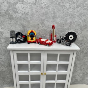 May include: Miniature rock and roll themed figurines including a microphone, a heart with "I Rock N Roll" text, a jukebox, a red convertible car, a red guitar, a motorcycle, and a vinyl record. The figurines are displayed on a white cabinet.