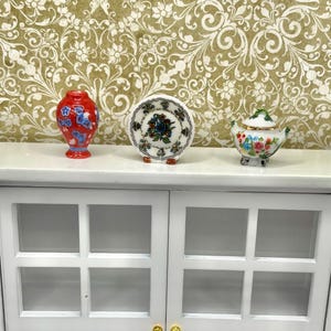 May include: Miniature ceramic decorative items on a white cabinet. The items include a red vase with blue floral designs, a small plate with a floral pattern, and a small teapot with a floral design. The cabinet has glass-paned doors.