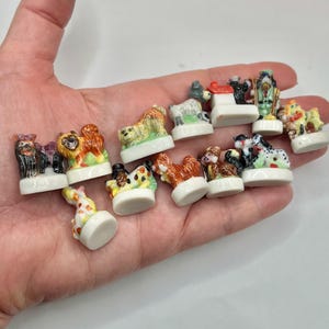 May include: A collection of small, colorful porcelain dog figurines. The figurines depict various dog breeds and are set on white bases. The dogs are painted with detailed features and vibrant colors, showcasing a variety of poses and styles.