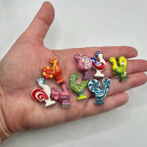 May include: A collection of small, colorful ceramic rooster figurines. The roosters are in various colors, including red, yellow, blue, and pink, with detailed designs. The figurines are held in a hand against a white background.