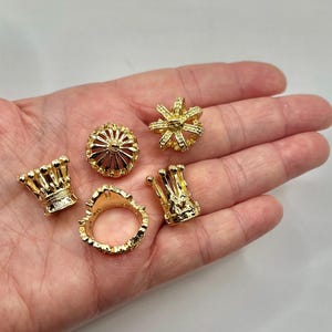 May include: Five small gold-colored metal crown-shaped charms or beads. Four of the pieces are solid, upright crown shapes with rounded points, while one piece is a circular ring band with a crown-shaped top, all featuring a metallic finish.