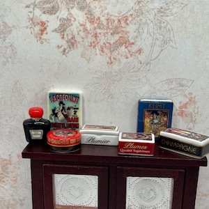 May include: Miniature antique perfume bottles and boxes on a dark wood cabinet. The collection includes a black bottle with a red stopper, and boxes with French text and vintage illustrations.