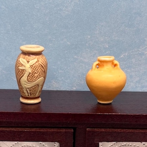 May include: Two miniature ceramic vases on a dark brown surface. One is tan with a deer design, the other is orange with handles. The vases are decorative and likely for a dollhouse or miniature collection.