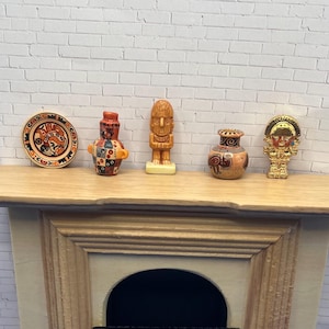 May include: Miniature ceramic figurines and a decorative plate are displayed on a wooden shelf. The collection includes a plate with a central design, a small vase, a carved wooden statue, and a gold-colored figure.