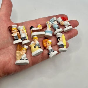 May include: A collection of nine small ceramic figurines depicting bakers in white uniforms with checkered pants. Each miniature figure is shown performing various tasks, such as holding bread, rolling dough, or carrying baking tools like rolling pins.