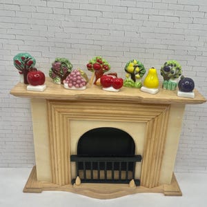 May include: A wooden fireplace mantel with a black grate and a collection of miniature ceramic fruit sculptures. The fruit includes apples, grapes, cherries, pears, and plums, each with vibrant colors and intricate details.