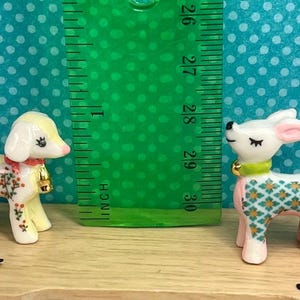 May include: Two ceramic figurines: a lamb with floral accents and a deer with a geometric pattern. The lamb has a red collar and a gold bell. The deer has a green collar and pink legs. A ruler shows the figurines are about 2.5 inches tall.