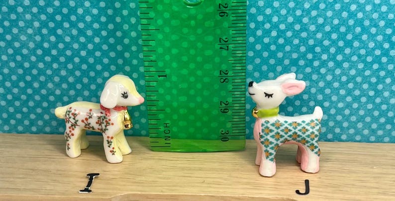 May include: Two ceramic figurines: a lamb with floral accents and a deer with a geometric pattern. The lamb has a red collar and a gold bell. The deer has a green collar and pink legs. A ruler shows the figurines are about 2.5 inches tall.