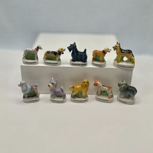 May include: A collection of ten miniature ceramic dog figurines, each standing on a small green base. The figures represent various breeds including a Dalmatian, Basset Hound, Scottish Terrier, Labrador, German Shepherd, Poodle, and others.