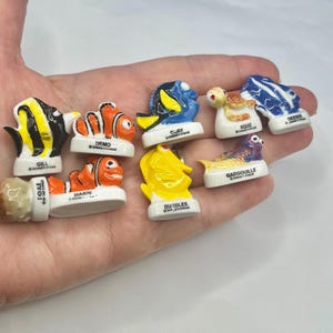 May include: A collection of small, colorful plastic figurines representing characters from Finding Nemo, each mounted on a white base with the character's name printed in black text, including Gill, Nemo, Marin, Dory, Bubbles, Squiz, Gargouille, and Debbie.