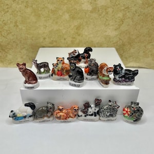 May include: A collection of fourteen small, hand-painted ceramic cat figurines arranged on a white display stand. Each miniature cat is posed differently, featuring various breeds and colors, with some cats interacting with small objects like balls.