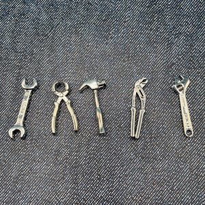 May include: A collection of miniature silver-colored tools, including a wrench, pliers, hammer, and adjustable wrench, arranged on a textured gray surface. The tools are detailed and appear to be made of metal.