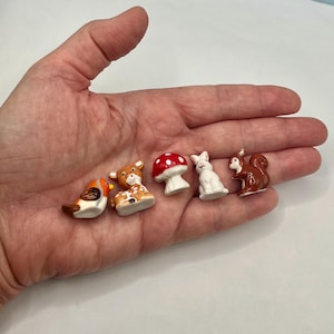 May include: A set of five miniature ceramic figurines including a small orange and white bird, a brown and white spotted fawn, a red and white spotted mushroom, a white rabbit, and a brown squirrel, all arranged in a row on a flat surface.