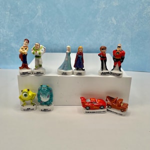 May include: A collection of Disney Pixar figurines, including characters from Toy Story, Frozen, The Incredibles, and Cars. The figurines are brightly colored and stand on white bases. The characters include Woody, Buzz Lightyear, Elsa, Anna, and Mr. Incredible.