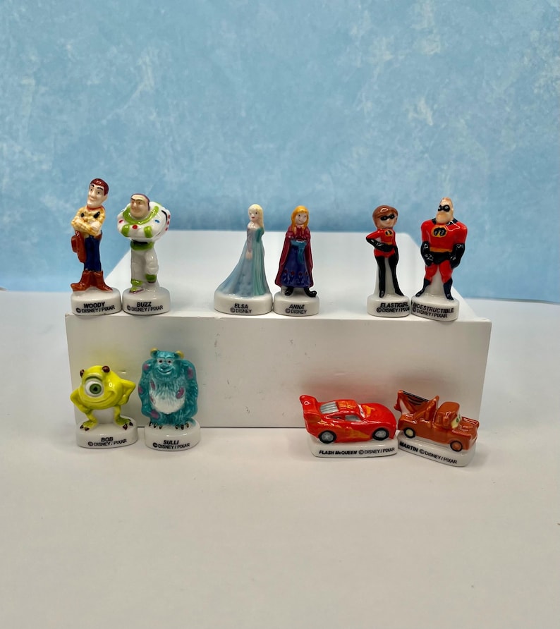 May include: A collection of Disney Pixar figurines, including characters from Toy Story, Frozen, The Incredibles, and Cars. The figurines are brightly colored and stand on white bases. The characters include Woody, Buzz Lightyear, Elsa, Anna, and Mr. Incredible.