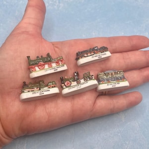 May include: Miniature ceramic train figurines in various colors, including green, red, and brown. Each train is mounted on a white base with text. The collection includes several different train models, showcasing intricate details and designs.