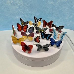 May include: A collection of colorful butterfly figurines displayed on a white rotating platform. The butterflies feature various colors, including red, yellow, blue, and black, with detailed wing patterns. The background is a light blue gradient.