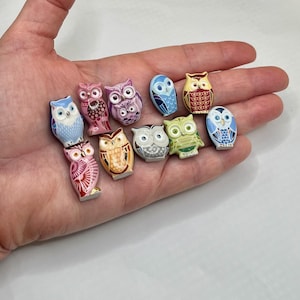 May include: A collection of ten small, flat-backed owl figurines in various colors and patterns, including shades of blue, pink, purple, yellow, green, and brown. Each owl features distinct decorative markings, large eyes, and a smooth, glossy finish.
