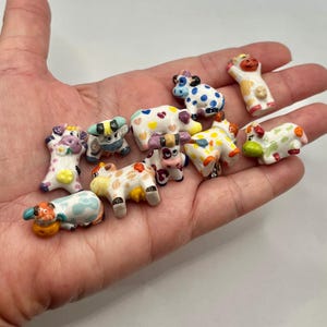 May include: A collection of small, hand-painted ceramic cow figurines. Each cow features unique patterns and colors, including polka dots, hearts, and floral designs. The figurines are in various poses, showcasing a whimsical and playful aesthetic.