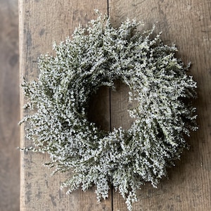 May include: A circular wreath composed of small, white flowers and green stems. The wreath is set against a rustic, wooden surface. This decorative wreath is suitable for indoor or outdoor use and adds a touch of natural beauty to any space.