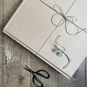 May include: A brown cardboard box tied with twine, adorned with a twine bow and sprigs of wheat. Black scissors rest on a wooden surface, suggesting a craft or gift presentation theme.