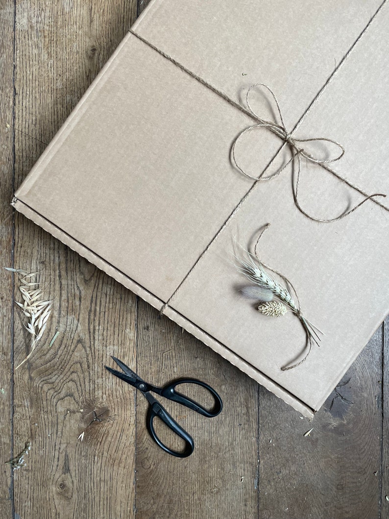 May include: A brown cardboard box tied with twine, adorned with a twine bow and sprigs of wheat. Black scissors rest on a wooden surface, suggesting a craft or gift presentation theme.