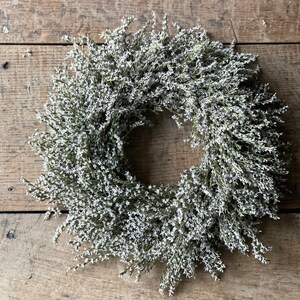 May include: A circular wreath composed of small, white flowers and green stems. The wreath is set against a rustic, weathered wooden background. The floral arrangement is dense and full, creating a natural, decorative element.