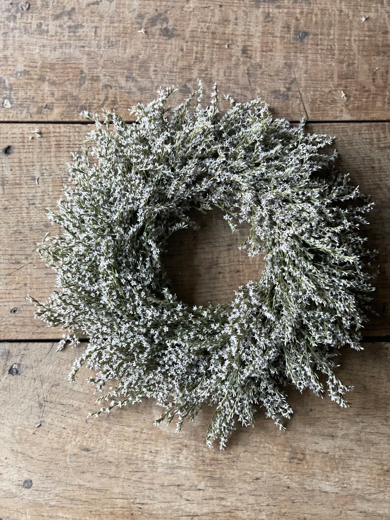 May include: A circular wreath composed of small, white flowers and green stems. The wreath is set against a rustic, weathered wooden background. The floral arrangement is dense and full, creating a natural, decorative element.