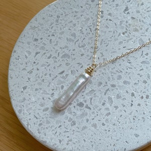 May include: A gold chain necklace with a long, white pearl pendant. The pearl is smooth and has a slight iridescent sheen. The necklace is displayed on a white, speckled surface.
