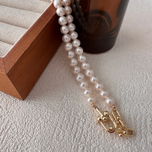 Peut inclure: Un collier de perles blanches à double rang avec un fermoir en or. Le collier est posé sur une surface texturée blanche.