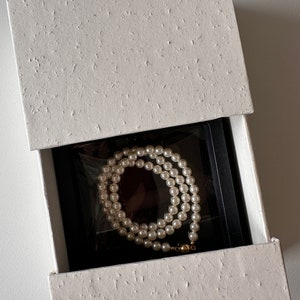 May include: A white pearl necklace with a gold clasp is displayed inside an open white textured gift box with a black interior. The necklace is neatly arranged, showcasing the lustrous pearls.