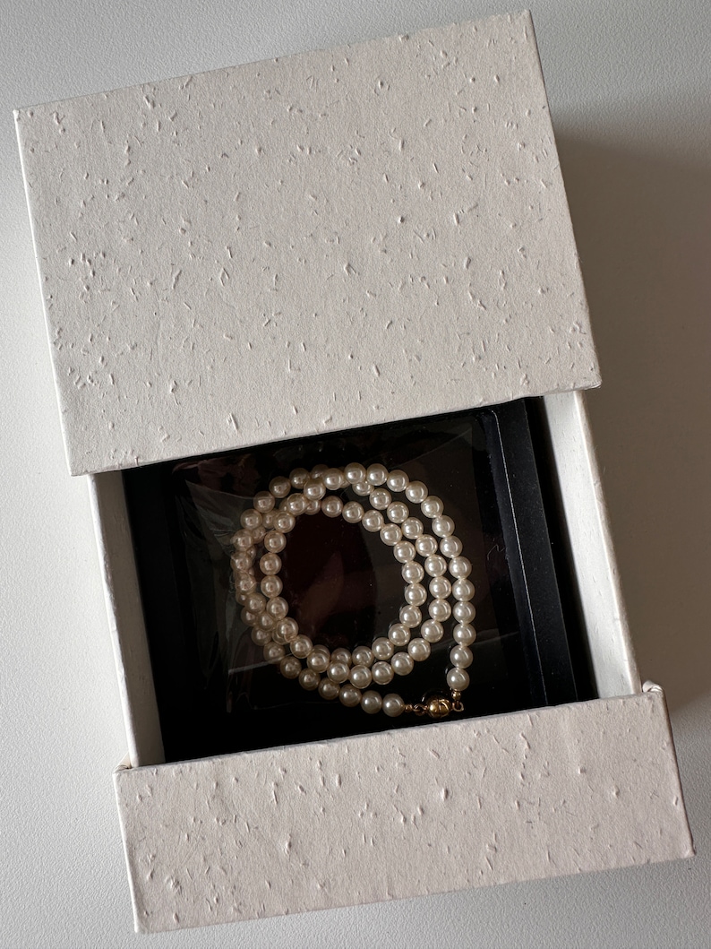 May include: A white pearl necklace with a gold clasp is displayed inside an open white textured gift box with a black interior. The necklace is neatly arranged, showcasing the lustrous pearls.