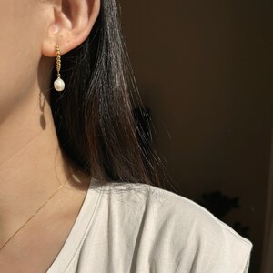 Puede incluir: Un pendiente de color dorado con un colgante de perla. El pendiente est&aacute; sujeto a la oreja con una peque&ntilde;a barra dorada texturizada. La perla es blanca y en forma de l&aacute;grima. El pendiente cuelga de la oreja.