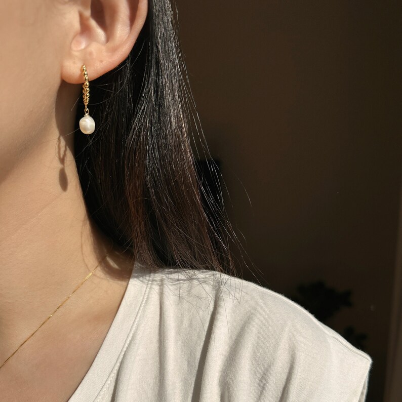 Puede incluir: Un pendiente de color dorado con un colgante de perla. El pendiente est&aacute; sujeto a la oreja con una peque&ntilde;a barra dorada texturizada. La perla es blanca y en forma de l&aacute;grima. El pendiente cuelga de la oreja.