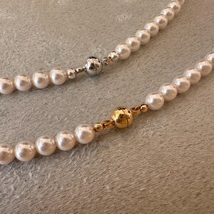 May include: Two pearl necklaces with magnetic clasps. One clasp is silver, the other is gold. The pearls are white and round. The necklaces are on a textured, light gray surface.