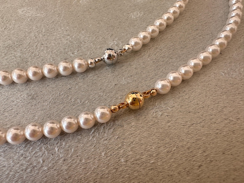 May include: Two pearl necklaces with magnetic clasps. One clasp is silver, the other is gold. The pearls are white and round. The necklaces are on a textured, light gray surface.