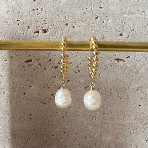 Puede incluir: Un par de pendientes de aro dorados con un dise&ntilde;o de cuentas. Cada pendiente presenta una perla blanca de forma irregular colgando. Los pendientes se muestran sobre un fondo de tono neutro.