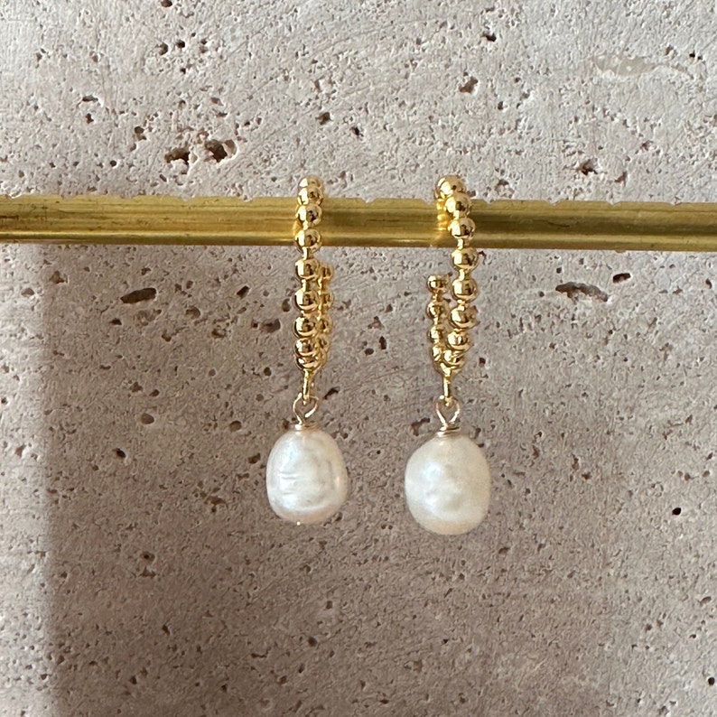 Puede incluir: Un par de pendientes de aro dorados con un dise&ntilde;o de cuentas. Cada pendiente presenta una perla blanca de forma irregular colgando. Los pendientes se muestran sobre un fondo de tono neutro.