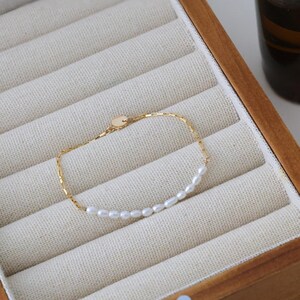 May include: A delicate gold chain bracelet featuring a single row of small white pearls. The bracelet is displayed in a wooden box with velvet lining.