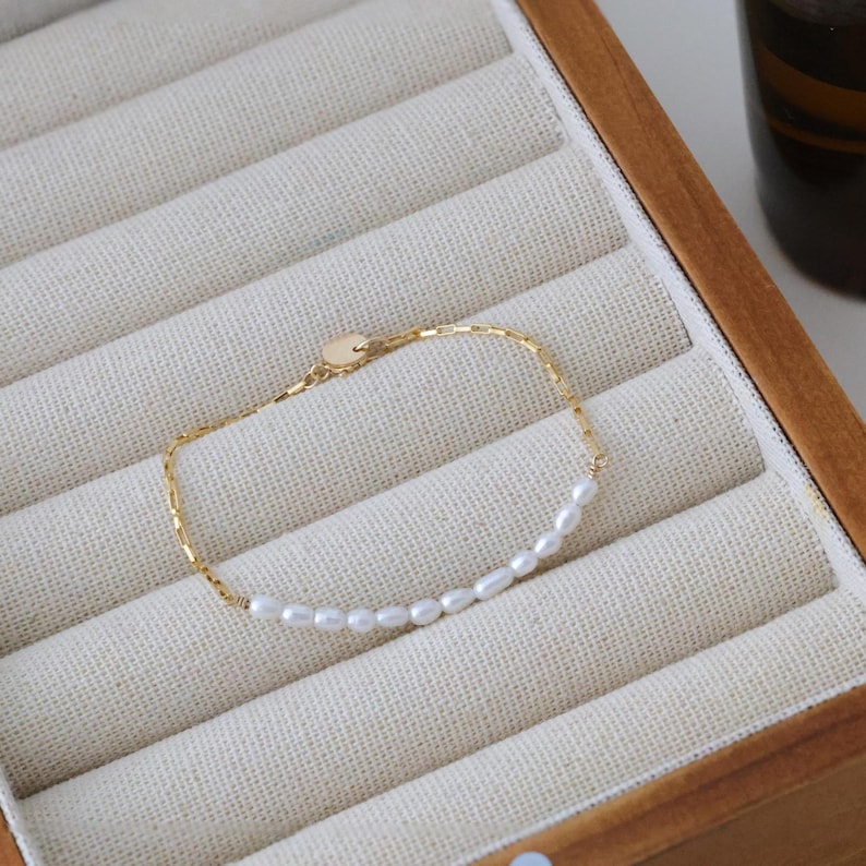 May include: A delicate gold chain bracelet featuring a single row of small white pearls. The bracelet is displayed in a wooden box with velvet lining.