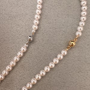 May include: A close-up of two strands of white pearl necklaces. One necklace has a silver clasp, and the other has a gold clasp. The pearls are round and evenly spaced, creating a classic and elegant look.