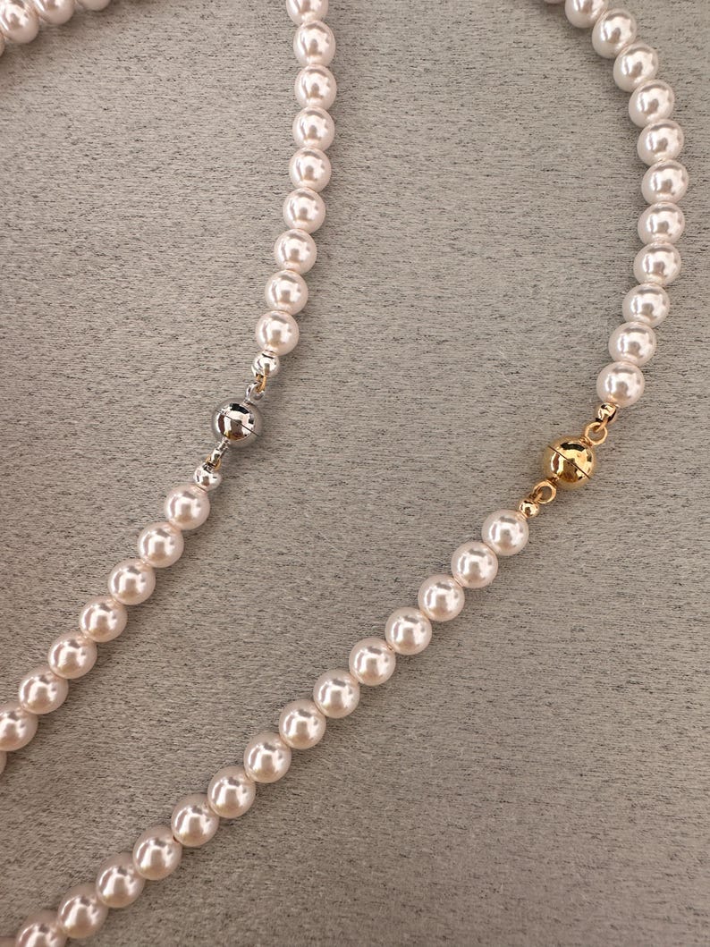 May include: A close-up of two strands of white pearl necklaces. One necklace has a silver clasp, and the other has a gold clasp. The pearls are round and evenly spaced, creating a classic and elegant look.