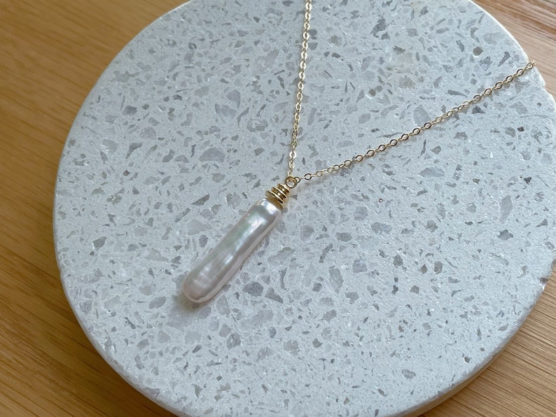May include: A gold chain necklace with a long, white pearl pendant. The pearl is smooth and has a slight iridescent sheen. The necklace is displayed on a white, speckled surface.
