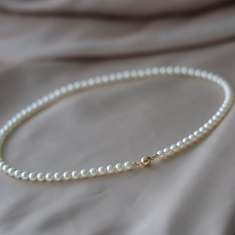 May include: A delicate pearl necklace with a gold clasp. The necklace features small, round, white pearls strung together. The clasp is a small, gold sphere. The necklace is displayed on a soft, neutral-toned background.