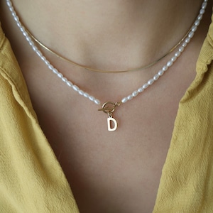 Può includere: Una collana con catena d'oro con un filo di perle e un ciondolo in oro a forma di lettera 'D'.